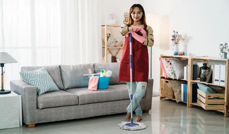 female housekeeper standing with keeping her arms on the mop in a bright home interior