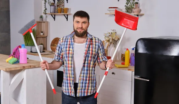 Young handsome bearded man in the kitchen wearing checkered shirt tries to deal with a dustpan